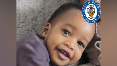 A close up of a baby with short black afro hair. He is leaning to the side and smiling at the camera. In the corner there is a West Midland Police logo