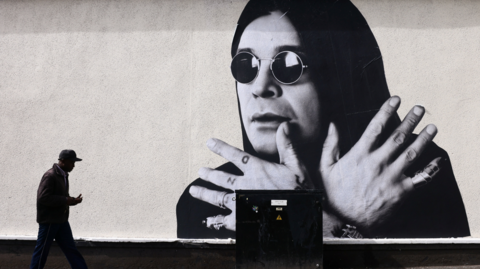 A man passes by a mural of Ozzy Osbourne on a white wall, depicting the former Black Sabbath frontman in his trademark round dark glasses, with hood up and his hands crossed in front of his throat.