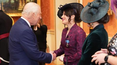 The King is shaking hands with Georgia after presenting her with the medal at Buckingham Palace. The King is wearing a navy blue suit and is looking directly at Georgia and taking with her. Georgia is wearing a maroon coloured dress with a hat and is talking to the King. Her sister Melissa is stood next to her wearing a green dress and watching on.