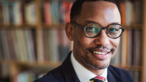 Mr Ibrahim Jalloh is stood to the right of the image looking at the camera. He has black hair, slight black and grey facial hair and circle frame glasses. He is smiling and standing against a full wooden book shelf while wearing a suit.