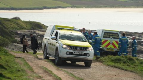 An image of a Coastguard truck on Greenaway Beach in Polzeath. There is one driving off the beach and another parked in the background. There are members coastguard in the usual blue uniform around the vehicles. 