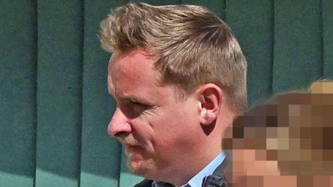 Connor McAreavey has short light brown hair, she is standing in front of a fence. 