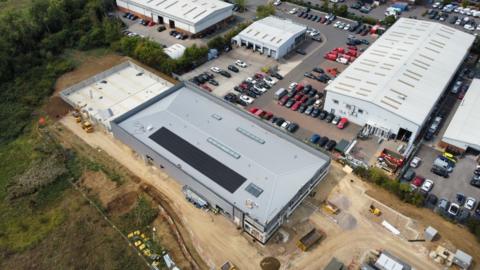 A drone shot of a large grey building with grass on one side, and sand around it. There is also large white buildings next to it and several parked cars.