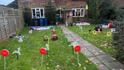 A house with a pathway through a large front garden covered with Christmas decorations and signs saying "Santa, stop here". Christmas lights are strewn across the front of the house.