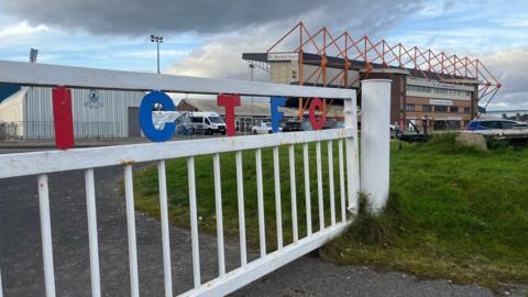 General view outside the home stadium of Inverness Caledonian Thistle. A white fence stands in the way of the stadium in the background. The fence has the letters I C T F C in red and blue at the top.