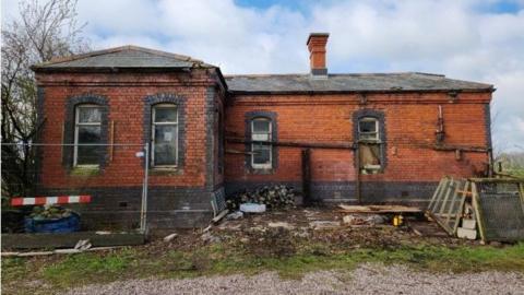 A large brick building with a chimney on the roof. The building is surrounded by metal gates and planks of wood.