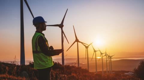 Silhouette of young male engineer holding laptop computer planning and working for the energy industry and standing beside a wind turbines farm power station at sunset time. 