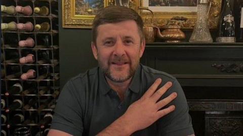 A man seated at a dining table in what appears to be an upscale or traditional-style restaurant or dining room. He is wearing a short-sleeved, dark-coloured polo shirt, and has brown hair with a beard.