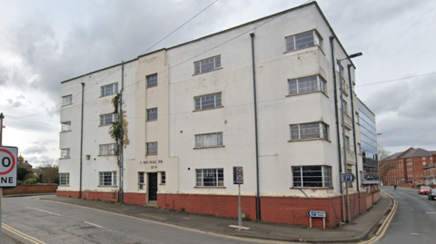 A large white four-storey building on the corner of a road. It has windows on each floor and a doorway. Above the doorway are the remains of sign that once read "John Haider building"