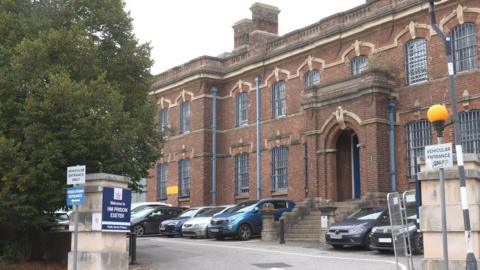 The entrance to and exterior of HMP Exeter on a grey day. On one of the stone entrance pillars is a sign reading: WELCOME TO HM PRISON EXETER and there are two signs reading: VEHICULAR ACCESS ONLY either side of the open entrance. There are cars parked next to the two-storey building which is red stone and looks historic as it has an ornate stone balustrade running around the roof line, many chimneys and pale stone features above the barred windows