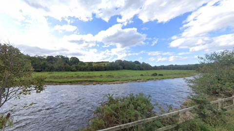 A Google Street View screenshot of the river. Taken from a nearby road, there is a flat, grassy bank on the other side, lined by large trees.