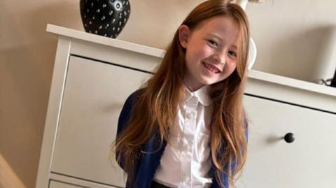 Nyla standing indoors in front of a white chest of drawers. She is smiling warmly at the camera with her hands held behind her back. She is wearing a school uniform consisting of a white collared shirt, a blue cardigan, a grey pleated skirt, grey tights, and black shoes. Her long, light brown hair falls neatly over her shoulders. On top of the chest of drawers behind her are decorative items, including a black vase with gold details and another vase containing dried foliage.