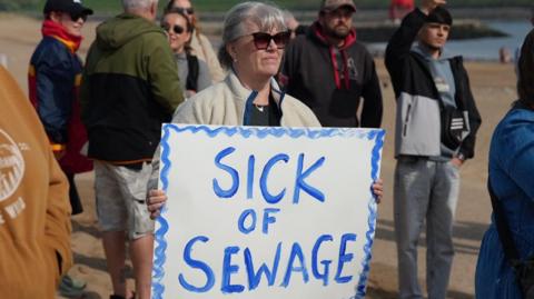 A woman holding a homemade sign on the beach. It is a white square sign with blue writing which says 'sick of sewage' with blue squiggly lines around the border. The woman has grey hair with a full fringe and is wearing large sunglasses. She is also wearing a white fleece. She has a straight face and is looking to the right of the camera. There is a group of people around her, some talking to each other and others looking in the same direction.