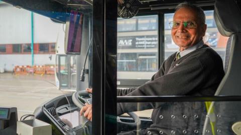 Amarjit Singh is sitting in the driver's seat of a bus, looking at the camera and smiling. He has his hands on the steering wheel and wears thin wire framed glasses with a blue collared shirt, tie and black long sleeved jumper.