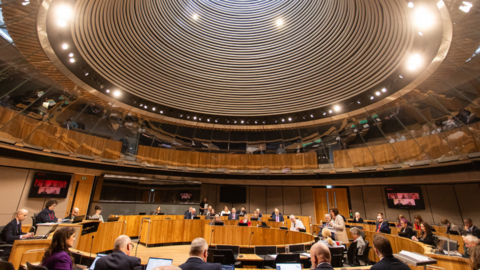 Senedd members in the recently expanded circular Senedd chamber with Eluned Morgan on her feet as other members listen while seated at their desks. The top of the picture is dominated by the large wooden funnel above the chamber.