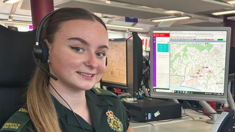 Lily Lambert at her desk at the South Central Ambulance Service Call Centre in Hampshire. She is wearing a green NHS uniform and there are computer screens at her desk, with maps on.
