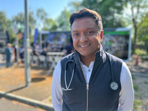 Navin Chandra, a consultant cardiologist, stands in front of a pop-up clinic in Slough. He is wearing a white shirt with a grey sleeveless jacket. He has a stethoscope over his shoulders and is smiling.  