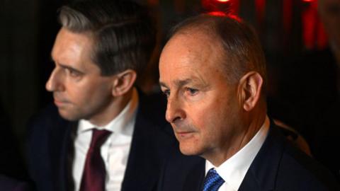 Two men are in frame. The man to the right is looking off-camera, he is bald with grey hair on the sides of his head. He wears a black suit white shirt and blue tie. The man on the right has grey/brown hair, he wears a navy suit and marron tie.