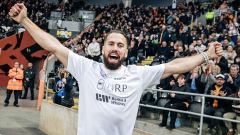 A man with slicked-back dark-brown hair and a beard stands with his arms aloft in front of a large crowd at a football stadium. He is wearing a white shirt with sponsor names on it. Behind him, football fans, sitting in rows of seats, applaud him.