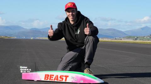 The image shows A man with a red baseball cap and a black hoodie, giving a double thumbs-up,  kneeling behind a remote control car on a long stretch of road.