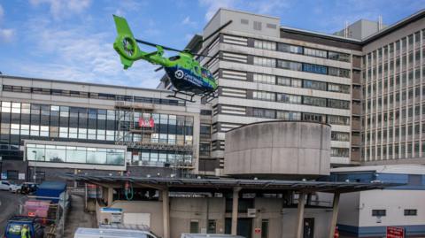 Great Western Air Ambulance's green and blue helicopter landing at a hospital. The building is several storeys high and has lots of vehicles parked outside.