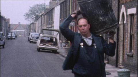 Singing binman Frank Clarke carrying a bin on his shoulder.