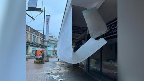 A large white stretch of plasterboard has fallen from a row of shops, and there is debris all over the ground. 