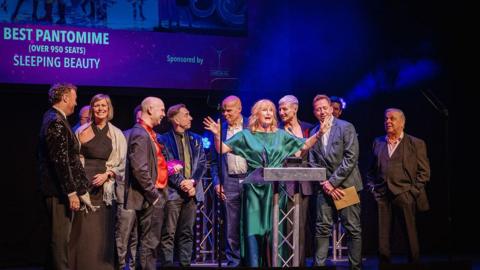 A group of smartly dressed men and women on a stage. A woman in a green dress, Karen Smallwood, stands at a podium giving a speech with her arms outstretched. In the background, a projector screen with the words 'best pantomime (over 950 seats) Sleeping Beauty