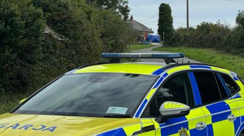A blue Garda road closed sign on a country road. Behind it is a police car and cordon. Further back is a building and a blue gazebo outside the building and officers. There are hedges and trees on either side of the road. The sky is cloudy.