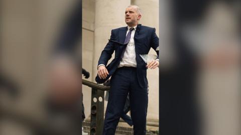 Actor Russell Tovey as Barclay in a navy blue suit. His tie is being swept by the wind and he is looking into the distance