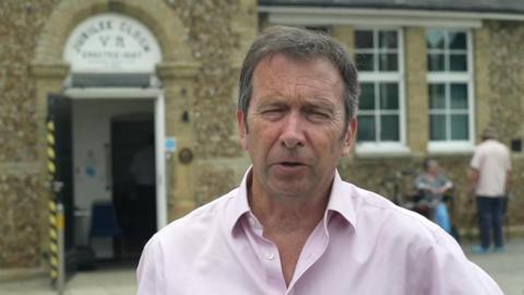 Alex Dunlop wears a pink shirt and looks to the camera. Behind is a stone building.