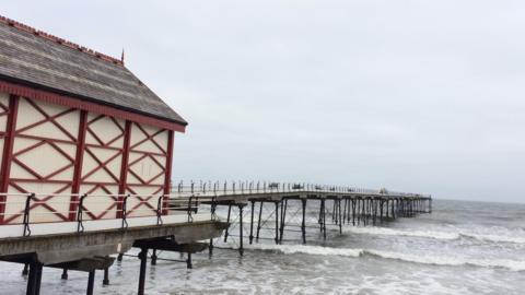 Sea waves crash into the bottom of Saltburn Pier on a gloomy day. The long, tall structure has benches on its deck. 