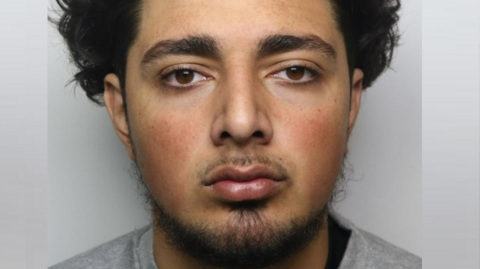 A mugshot of Ryan Willicombe, wearing a grey, prison issue sweatshirt, and has dark, curly hair and a short moustache and beard 