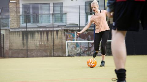 A woman with white blonde hair in a ponytail and a peach T-shirt with black leggings is kicking a ball on an outdoor football pitch. Someone blurred in the foreground is standing in front of her.
