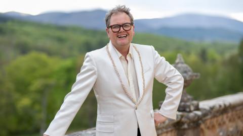 Alan Carr standing outside the Traitors castle in the Highlands