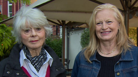 Diana Parkes has short white hair and is wearing a black coat, a pink jumper and a white, buttoned-up shirt and a black scarf. Hetti Barkworth-Nanton is stood next to her. She is smiling and has shoulder length blonde hair. She is wearing a denim jacket and black shirt and a gold necklace around her neck