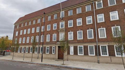 A large red brick building, with a symmetrical design, comprises four storeys and a large wooden double door.