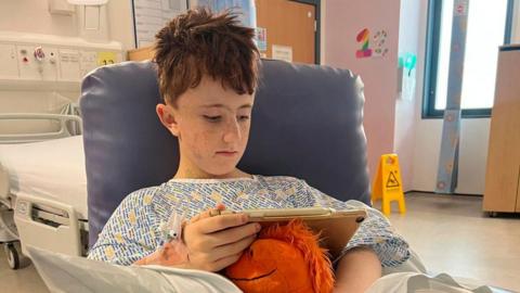 A young boy sitting his a chair wearing a hospital gown, he is looking down at an ipad and holding an orange soft toy,
