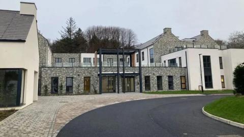 The front of Summerhill View care home. A large building predominantly featuring grey brickwork with some white walls. A section in the middle shows balconies at the front of rooms on the second floor. Brick paving and a grassed area lie at either side of a newly tarmacked curving road in front of the building. 