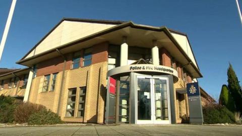 The outside of Police and Fire headquarters in Portishead, Somerset. The building has a large circular entrance with a revolving door, which has the words "Police and Fire Headquarters" above it. The building has contrasting light and dark brick work and a light-coloured, pointed roof. The picture is taken from a very low angle, almost from the ground. 