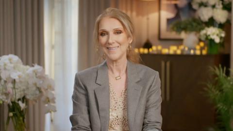 Céline Dion sitting on a chair with flowers behind her, smiling and wearing a grey blazer with a cream top