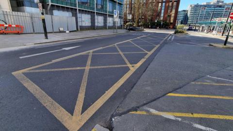 A long yellow box marking spreads along a junction. No vehicles are seen on the road with a number of large buildings in the background.