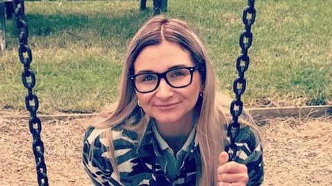 Isobella Knight with long blonde hair wearing large dark brown glasses. She is wearing a black and gold top and is holding onto the chain of a swing in a park. There is a dried grass landing area behind her and green grass beyond that.
