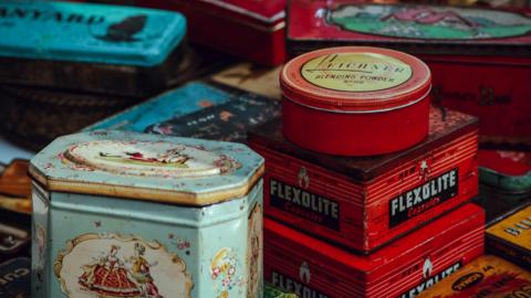 An assortment of old household goods tins, partially rusted. A pale blue tin is in the foreground, with a picture of a regal looking lady and squire dressed in formal clothing from centuries ago. A red tin labelled Flexolite capsules sits next to it, with another red one for Leichner blending powder on top.