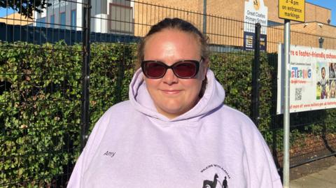 A woman with brown hair, tied back, wearing sunglasses and a light-pink hoodie is standing in front of a modern brick school building, which is surrounded by a black wire fence and green hedge. A yellow parking sign reads: "No stopping Mon - Fri, 8-9am, 3-4 pm on entrance markings". Another sign advertises fostering services.