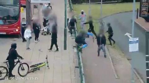A group of masked young people at a bus stop. One appears to be carrying in a weapon. 