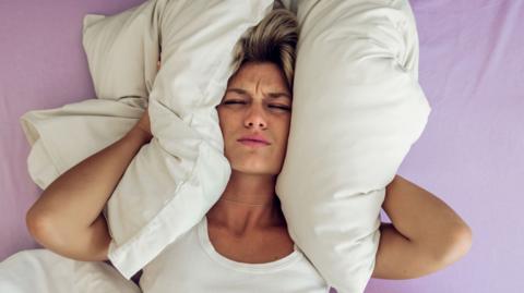 Woman in white top lying in bed with pillows pushed to her ears to drown out noise