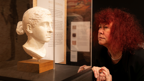 A carved stone bust is displayed inside a glass case in a museum. The bust shows a head with detailed hair tied at the back. Behind the case, exhibition panels with text and images are visible. A person with curly hair stands close to the display, leaning forward with hands clasped and looking at the sculpture.