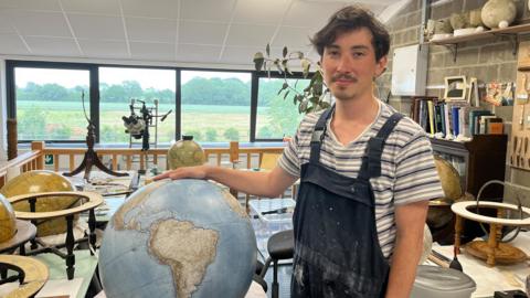 A man stands in a bright workshop beside a large, detailed globe mounted on a wooden base, with one hand resting on it. The workspace is filled with tools, brushes, and circular frames used in globe-making, neatly arranged on a wall and surrounding tables. Several partially completed globes and spherical models are visible, indicating an active craft or restoration process. Large windows in the background let in natural light and reveal a green outdoor landscape.