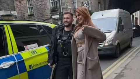 One of the police officers is standing on the road in between his police car and Tallia Storm. She is wearing a long brown coat. There is the arch of a building in the Cowgate in the background.